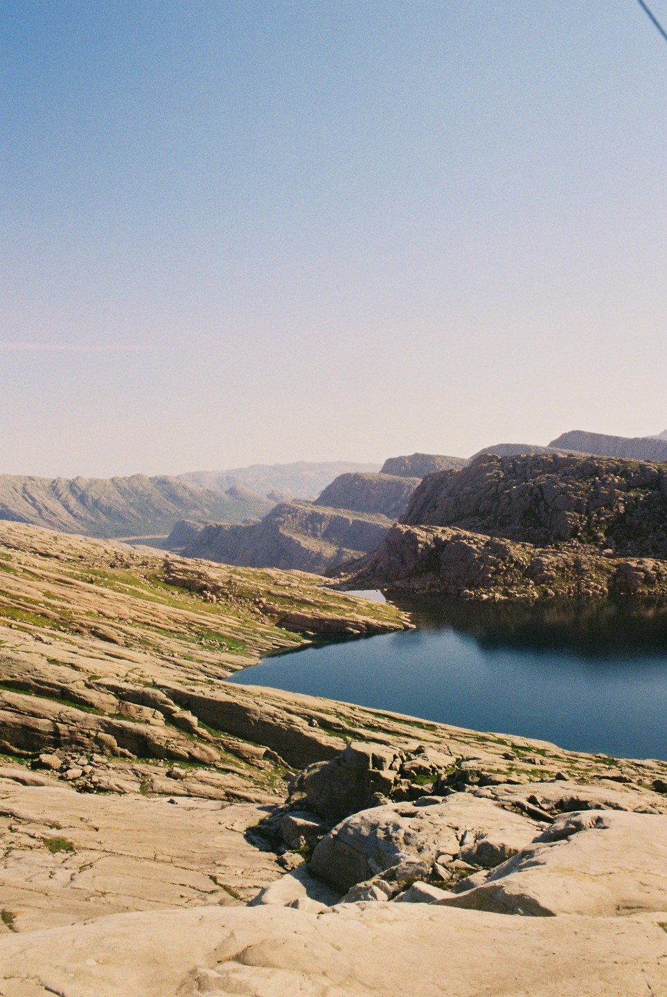 A body of water is nestled among rocky, stepped mountains under a clear sky.