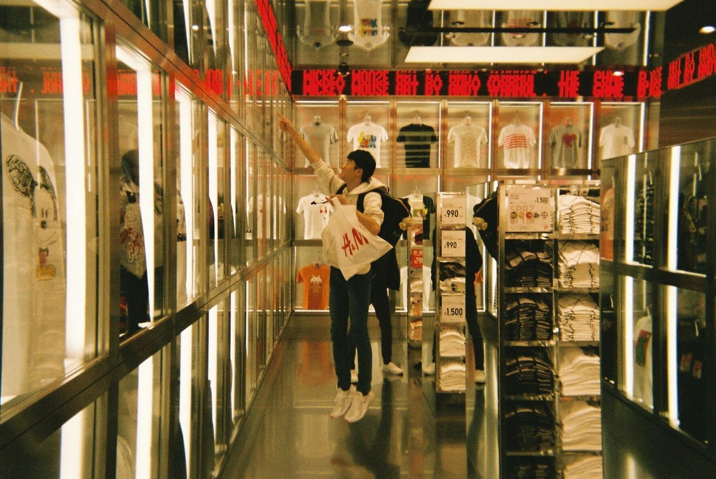 A man with a backpack and a shopping bag reaches for a t-shirt in a brightly lit clothing store.