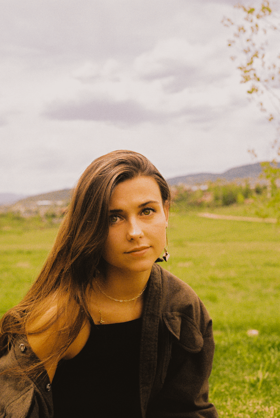 A woman with long hair and a dark jacket looks directly at the viewer in a grassy field with hills in the background.