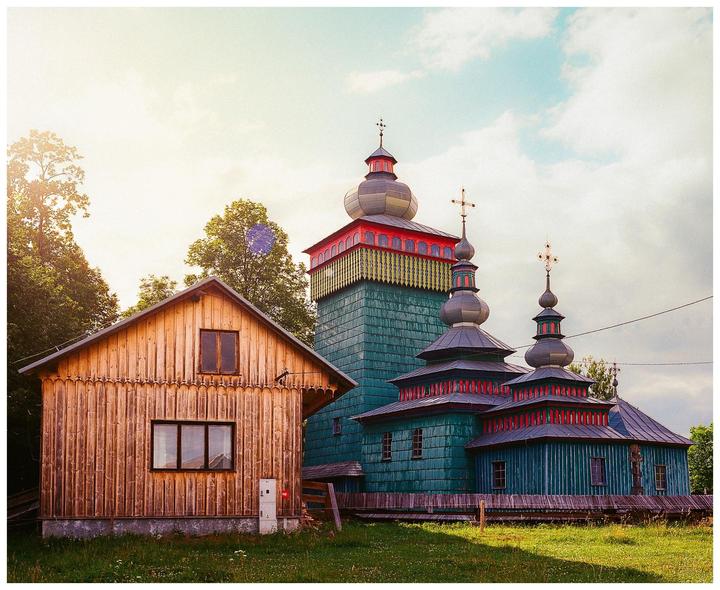 A wooden church with multiple domes and a cross on each dome stands next to a wooden house in a grassy field.