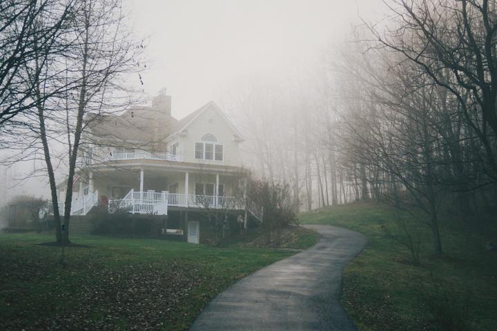 A large house with a porch and white railings sits on a hill next to a winding path and a forest, all shrouded in fog.