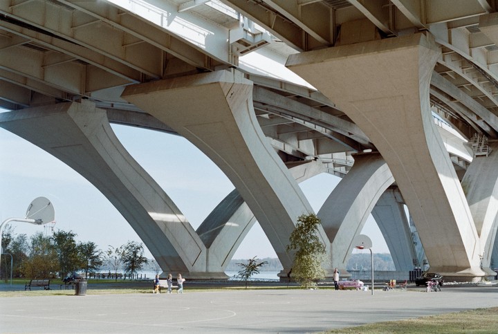 A wide shot shows the underside of a large concrete bridge with multiple arches, under which a basketball court and several people are visible.