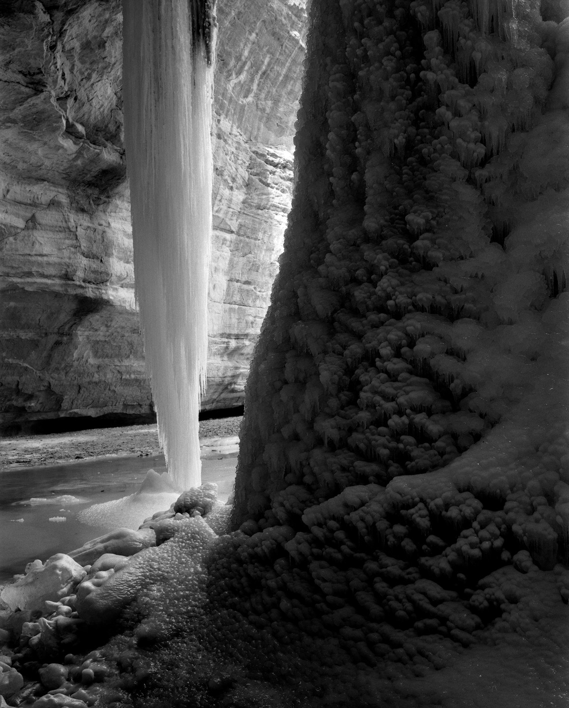 A large column of ice stands next to a textured ice formation in a rocky canyon with a stream.
