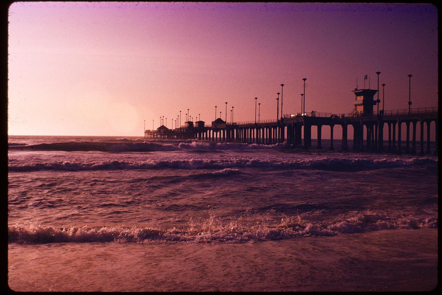 Waves crash on a beach with a pier extending into the ocean under a pink sky.