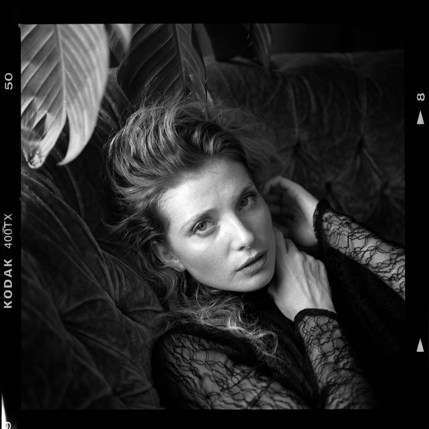 A woman with wavy hair and a lace top is reclining on a couch with large leaves around her.