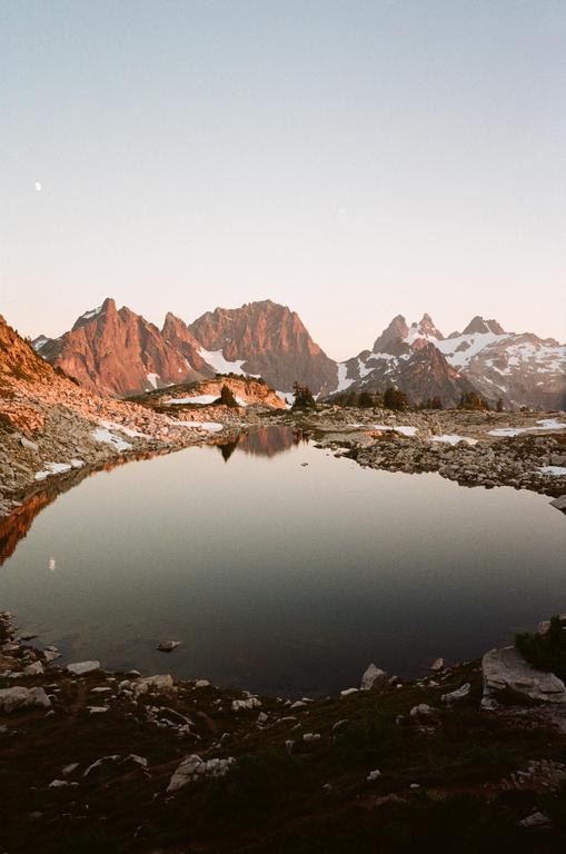 A calm lake reflects mountains with snow and a moon in the sky during golden hour.