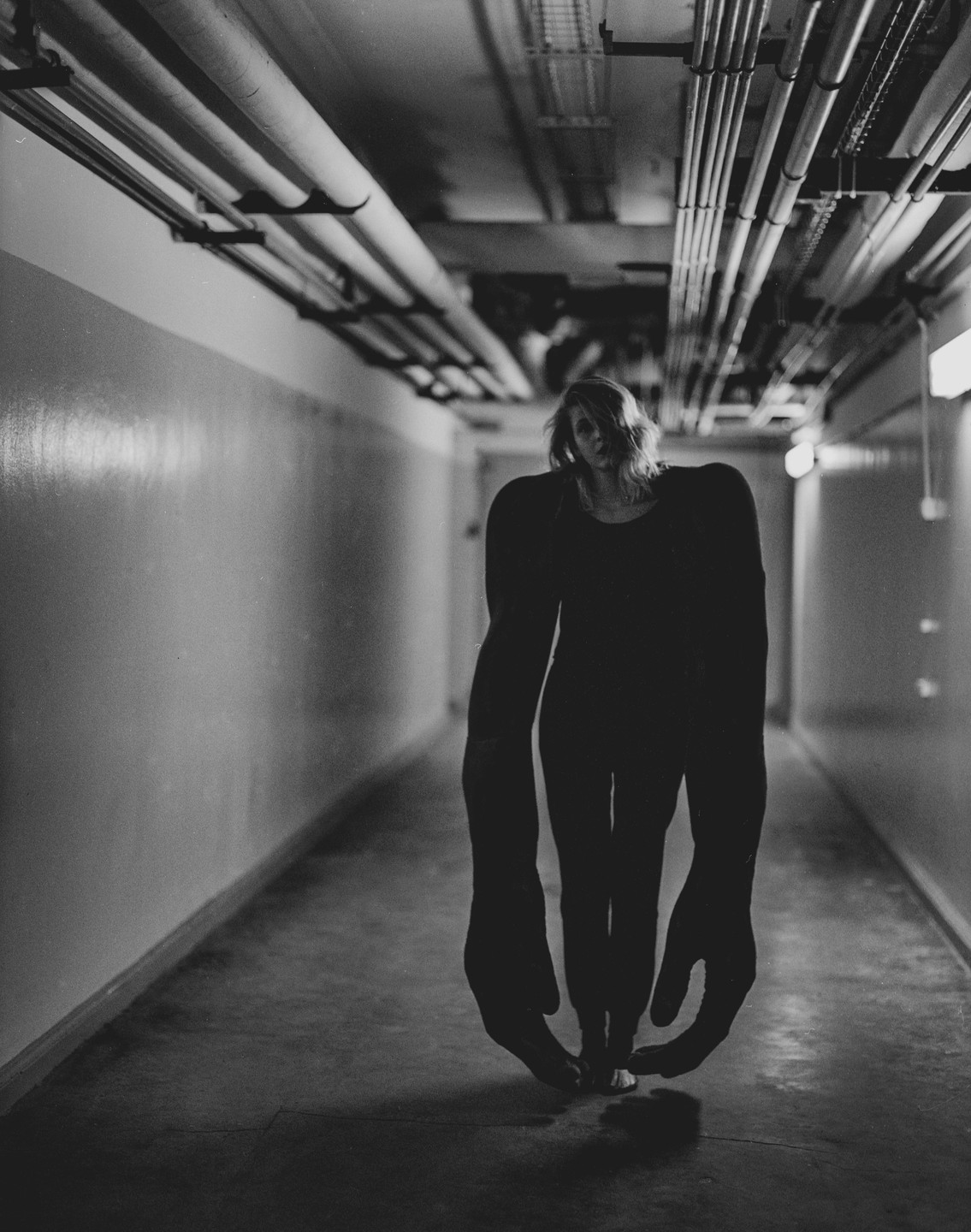 A woman with exaggeratedly long arms and large hands walks barefoot down a dimly lit industrial hallway.