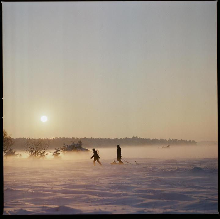 Two people walk across a snowy field pulling a sled as the sun sets behind a foggy forest.