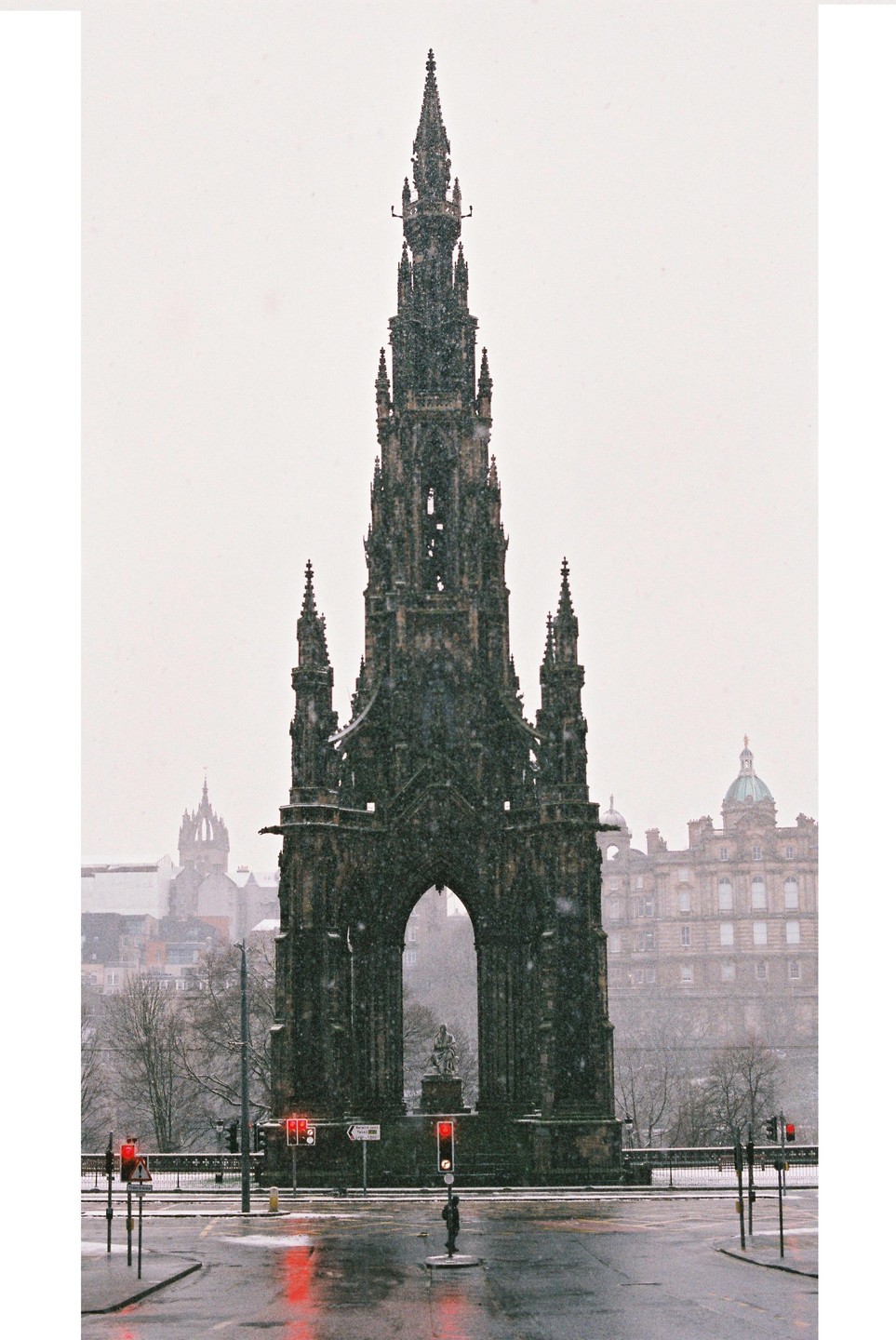 A tall, dark gothic monument stands in the middle of a snowy street with red traffic lights reflecting on the wet ground.