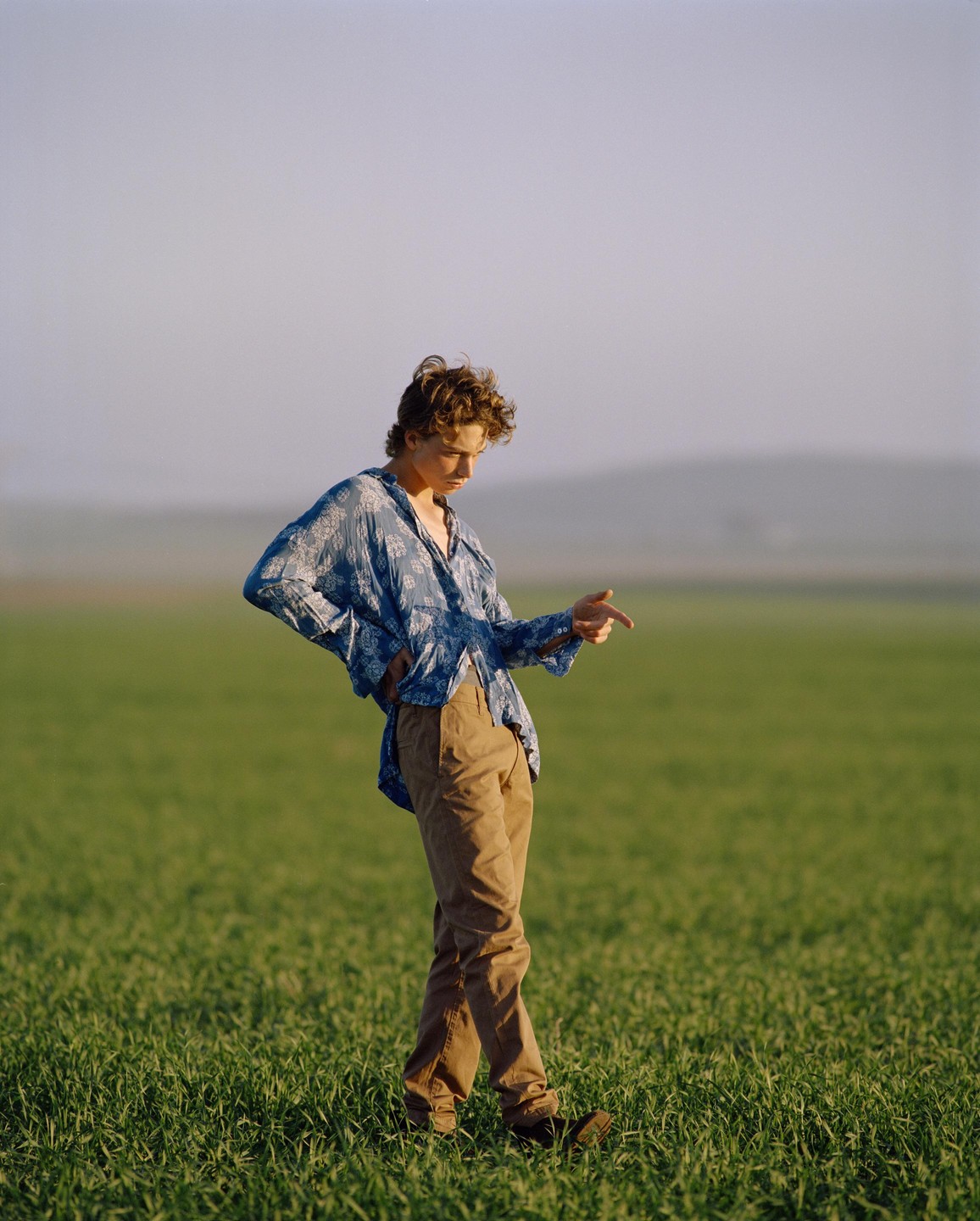 A young man in a blue shirt and brown pants stands in a field of green grass, looking down and pointing.