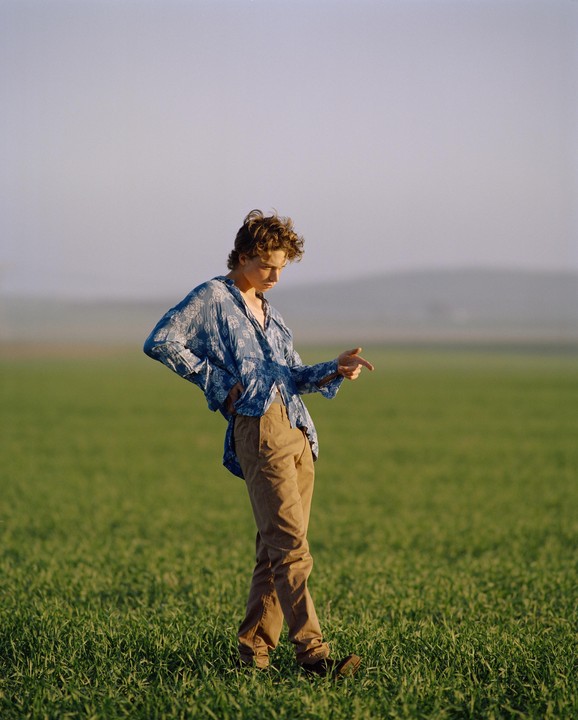 A young man in a blue shirt and brown pants stands in a field of green grass, looking down and pointing.