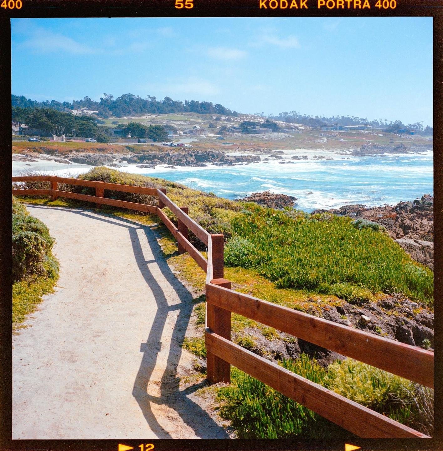 A dirt path with a wooden fence runs along a coastline with a bright blue ocean and green vegetation under a clear sky.
