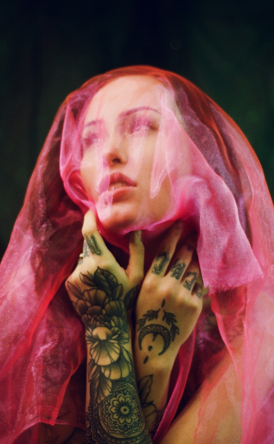 A woman with tattooed arms and hands is partially obscured by a sheer pink veil.