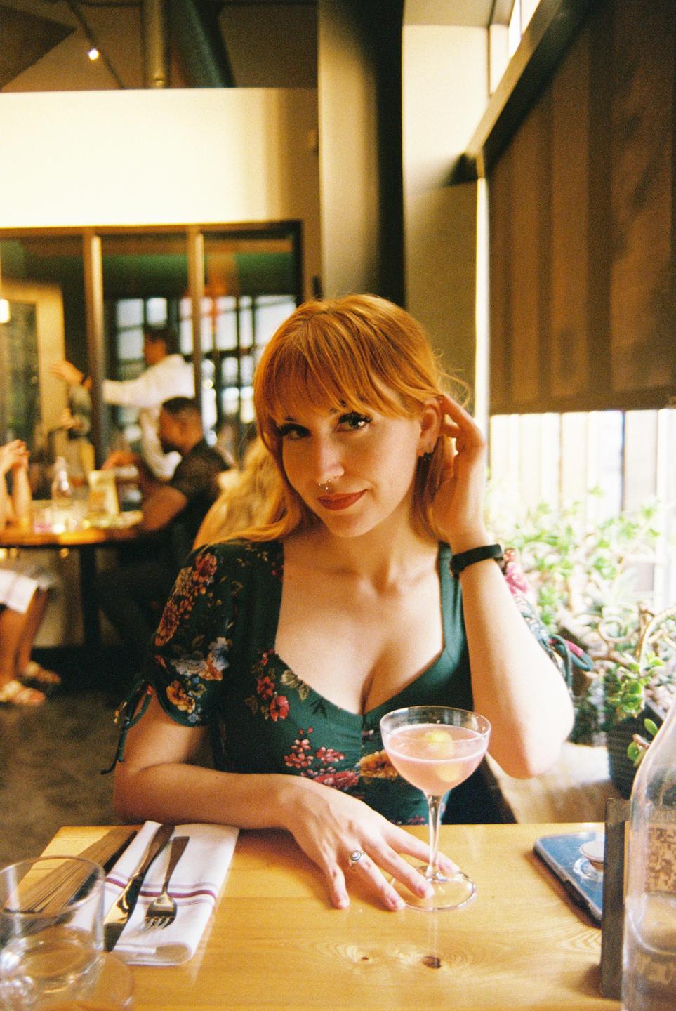A woman with red hair sits at a table in a restaurant with a pink cocktail.