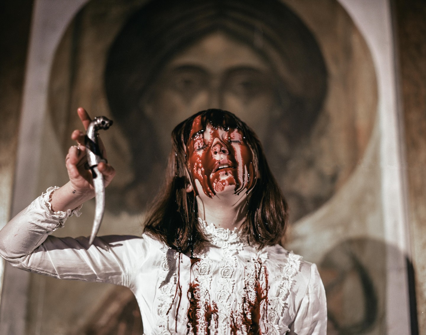 A woman with blood on her face and white dress holds a dagger in front of a painting.