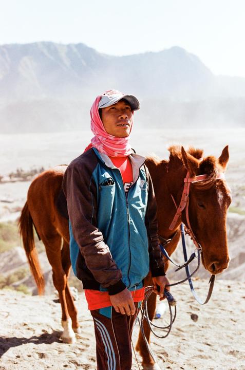 A man wearing a pink head covering and a blue jacket stands next to a horse in a mountainous, arid landscape.