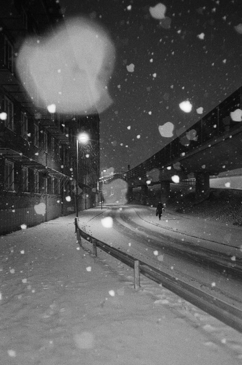 A person walks down a snow-covered street at night, illuminated by a street light, with a bridge overhead.