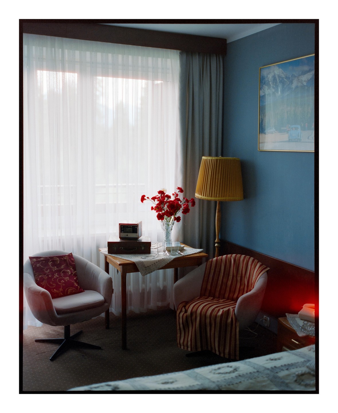 A hotel room features two armchairs, a small table with a vase of red flowers, and a floor lamp.