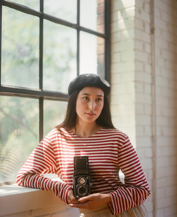 A woman wearing a striped shirt and beret holds a camera while standing next to a window.