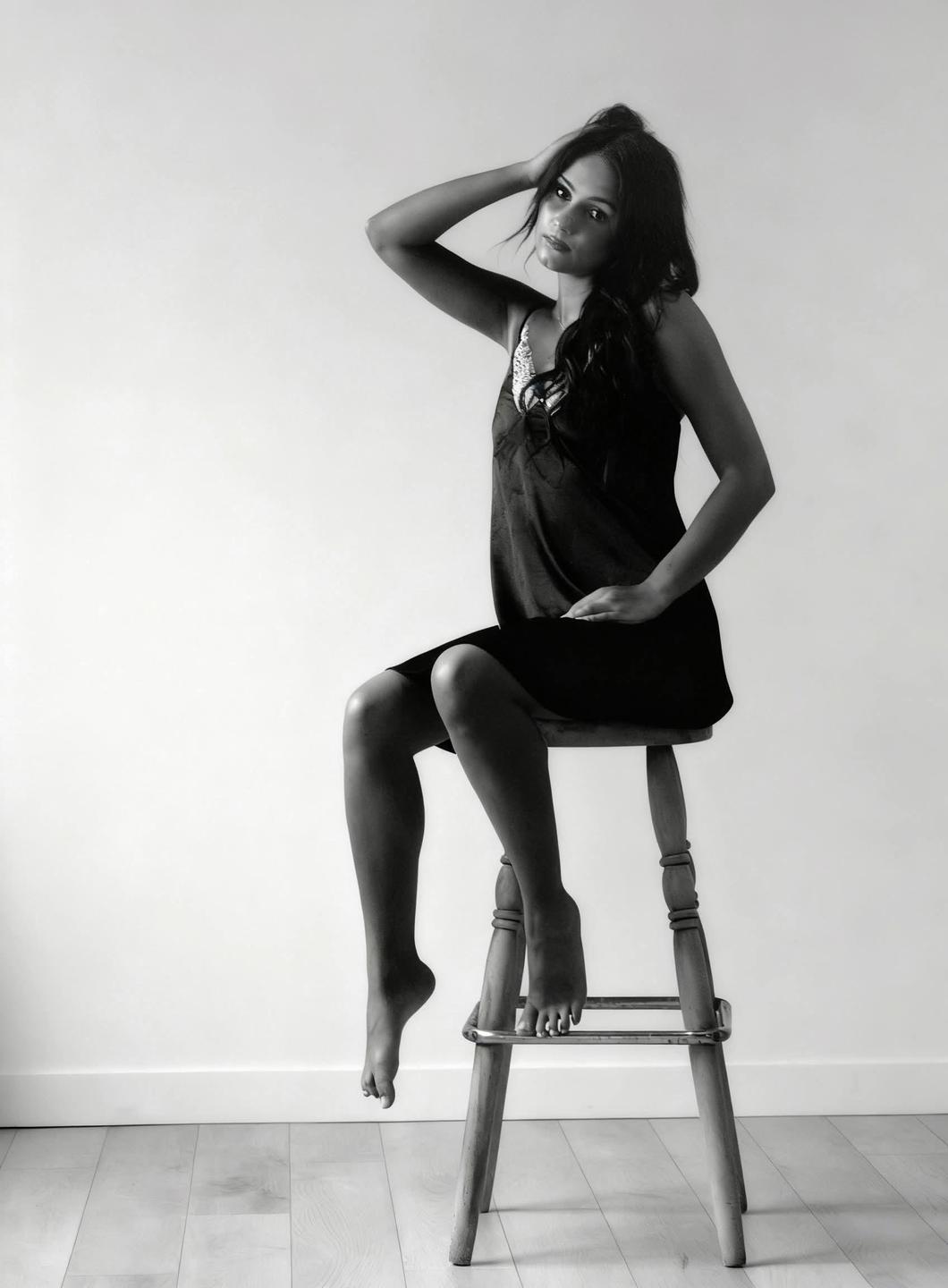 A woman in a dress sits on a stool with one arm raised to her head.
