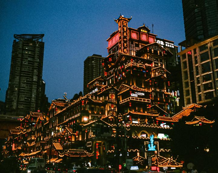 A multi-story building with traditional Chinese architecture is brightly lit at night, surrounded by modern skyscrapers.