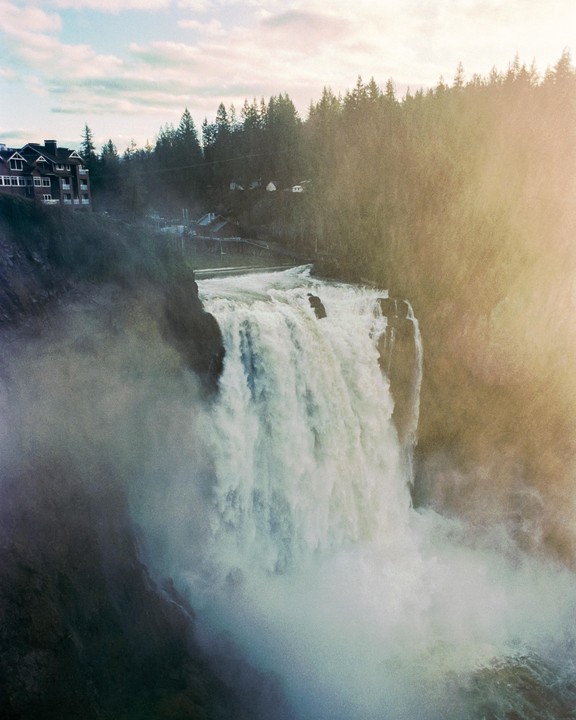 A large waterfall cascades down a cliff next to a building and a forest.