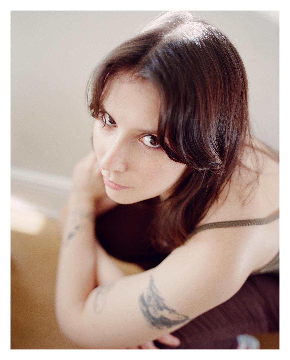 A woman with dark hair and tattoos on her arms looks up at the viewer.