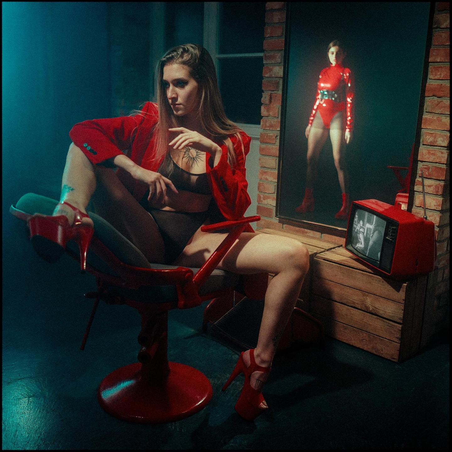 A woman in a red jacket and high heels sits in a chair, with her reflection visible in a mirror and a red television set nearby.