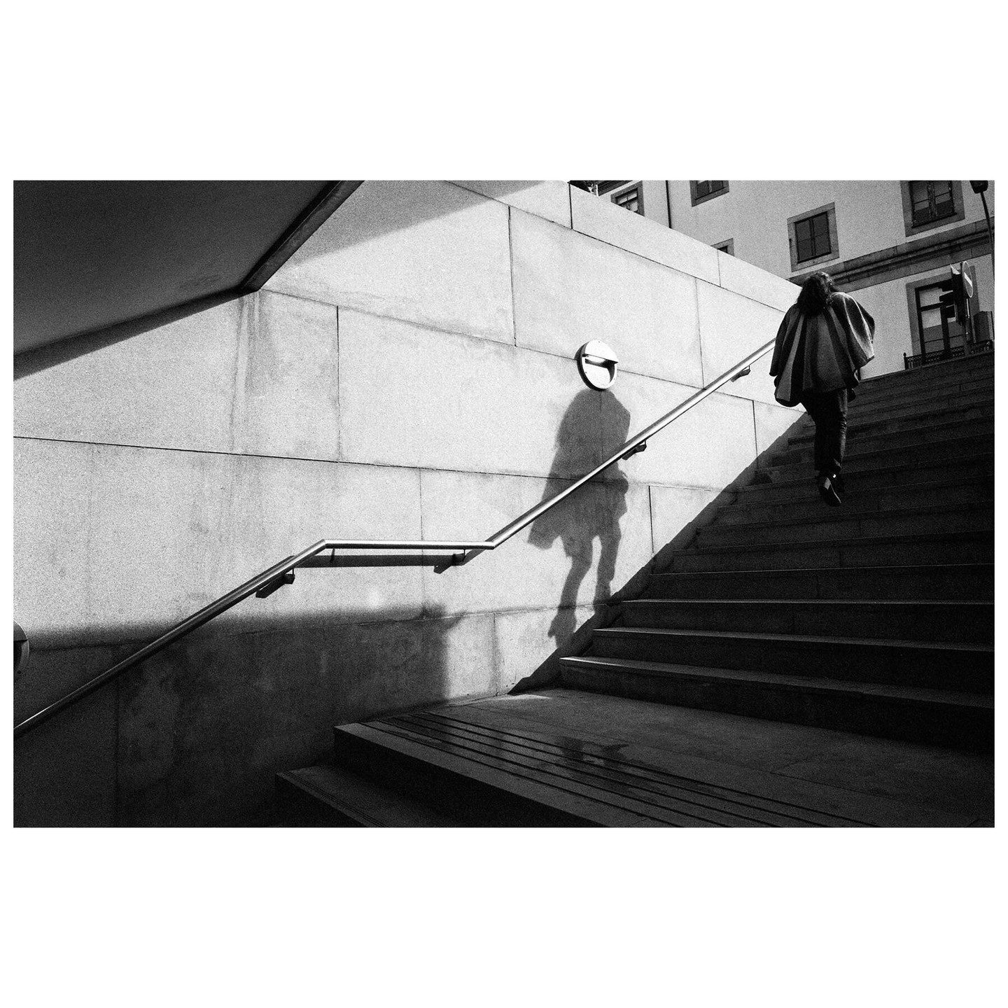 A person walks up a set of outdoor stairs next to a wall with a long shadow cast on it.