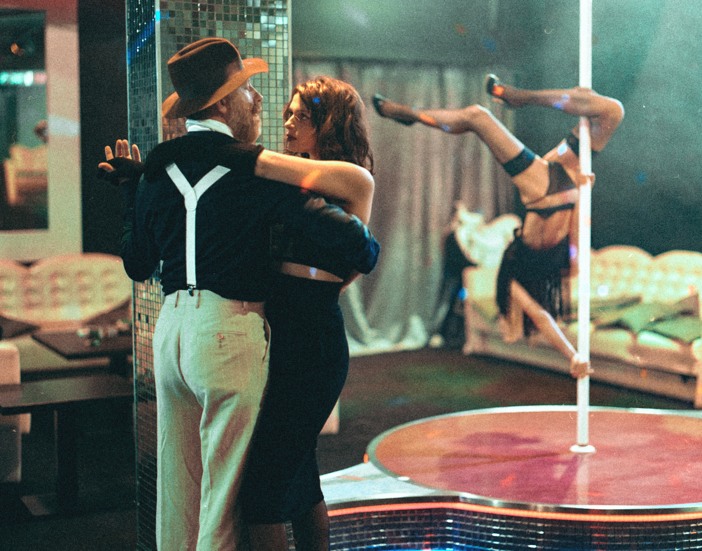 A man in a hat and suspenders dances with a woman in a dark dress while another woman pole dances in the background.