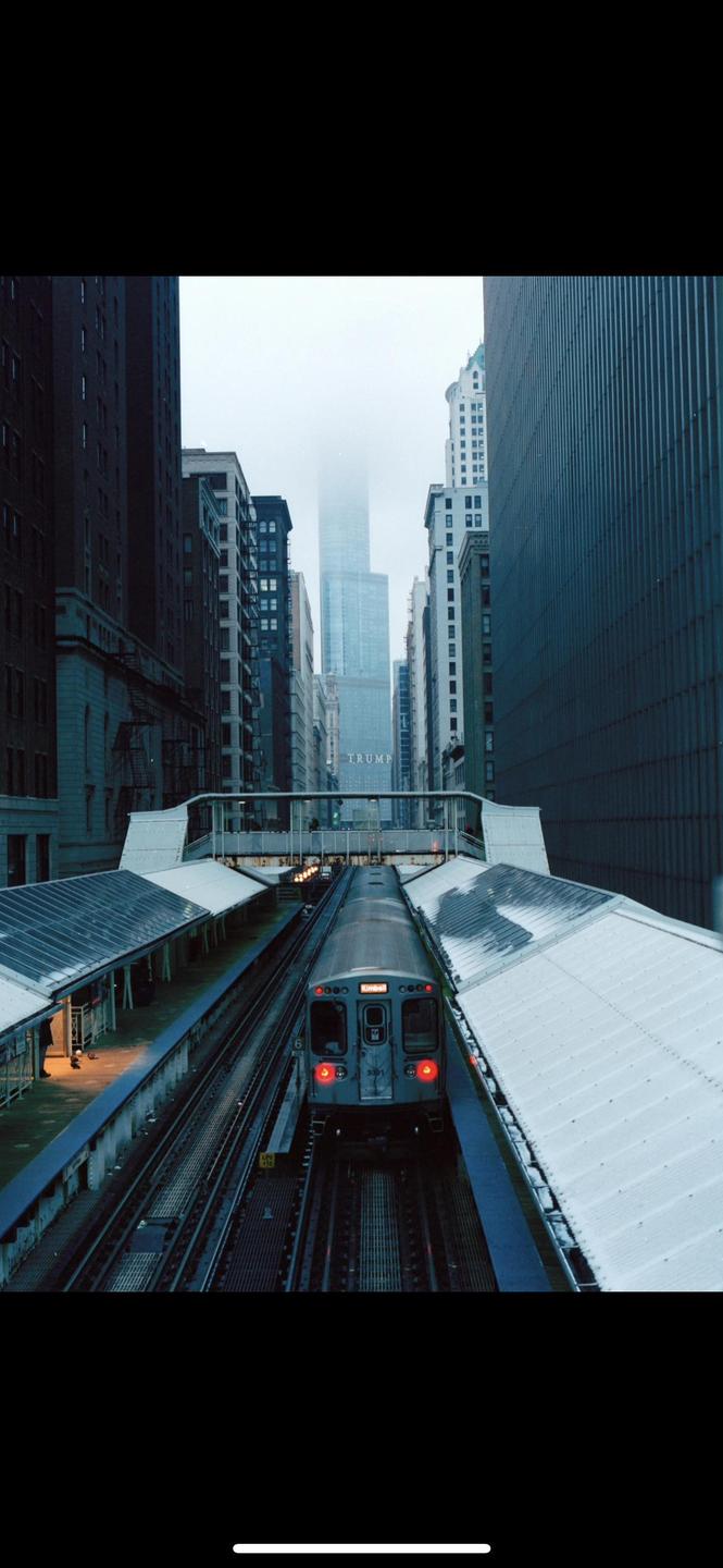 A train is visible on tracks between two platforms with tall buildings in the background, one of which is partially obscured by fog.