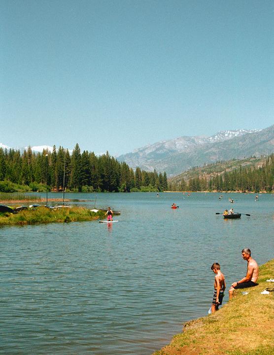 A man and a boy sit by the edge of a lake with mountains and a forest in the background.