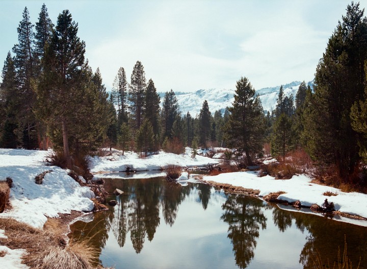 A river flows through a snowy forest with mountains in the background.