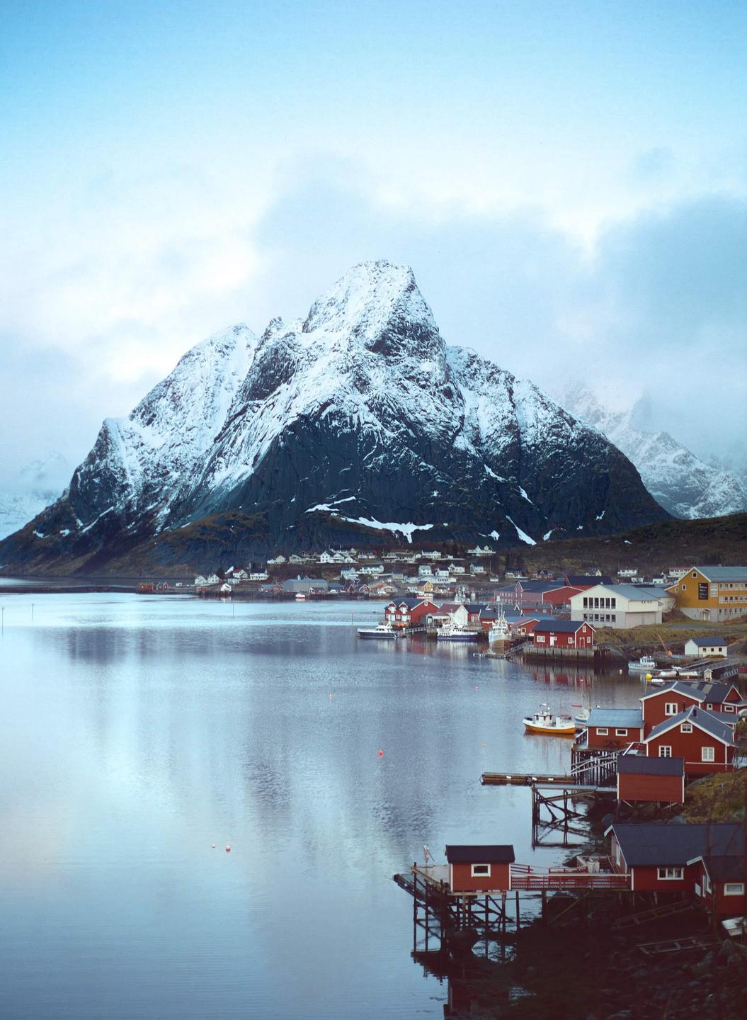 A village with red and yellow houses sits on the shore of a body of water, with a large snow-capped mountain in the background.