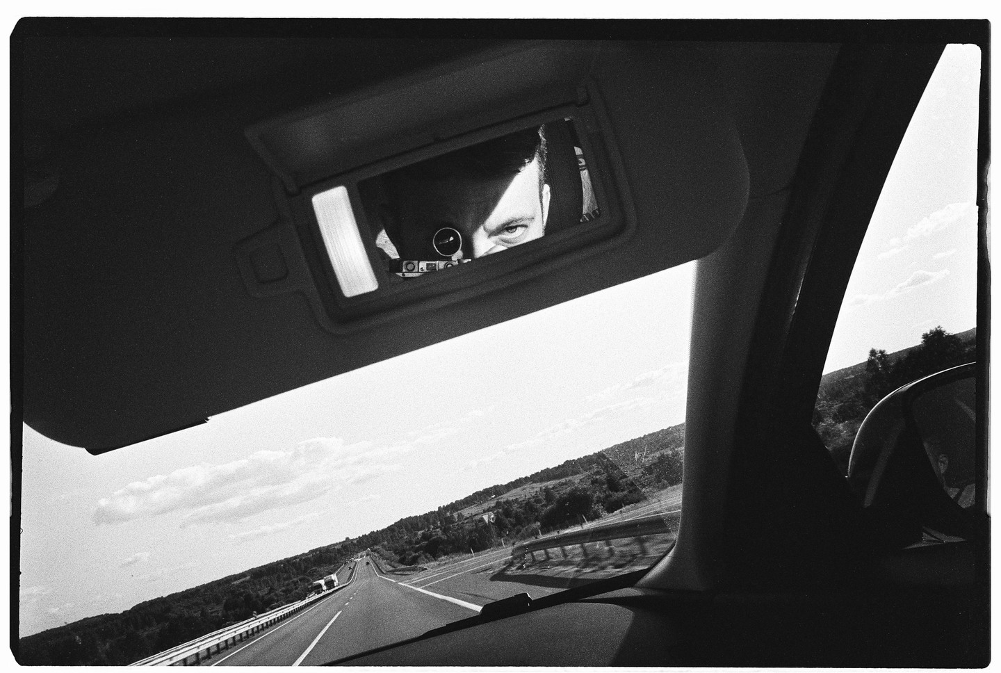 A man's eyes are visible in a car's sun visor mirror, with a highway stretching into the distance outside the car.