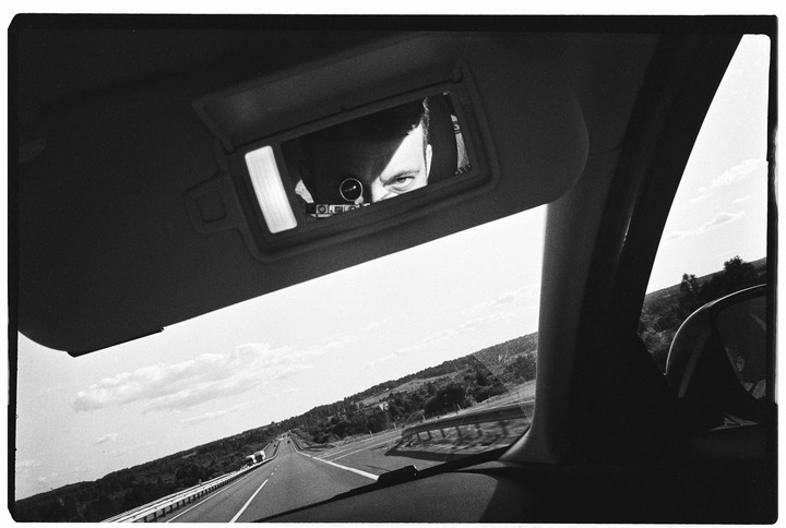 A man's eyes are visible in a car's sun visor mirror, with a highway stretching into the distance outside the car.