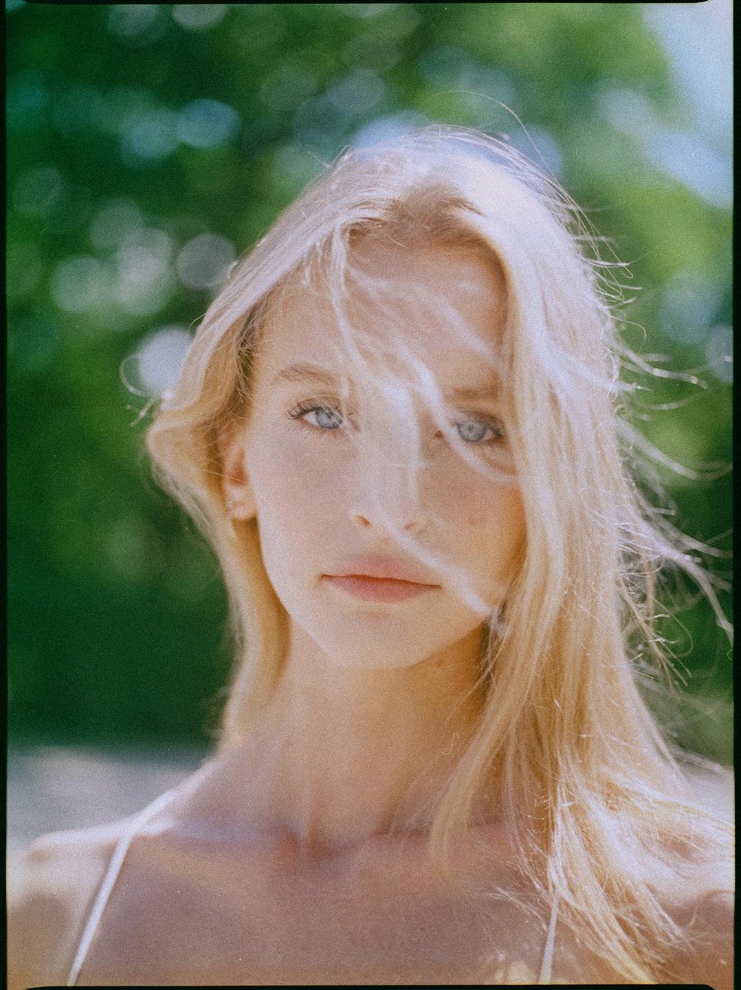 A woman with blonde hair and blue eyes looks directly at the viewer, with her hair blowing across her face.