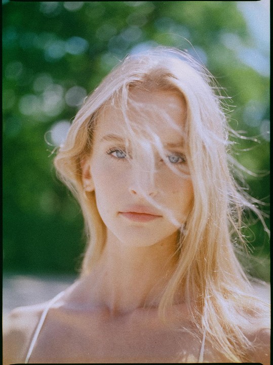 A woman with blonde hair and blue eyes looks directly at the viewer, with her hair blowing across her face.