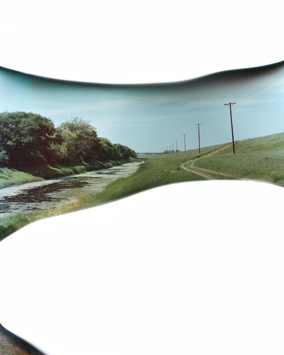 A narrow river with trees on its bank runs alongside a grassy hill with a dirt path and telephone poles.