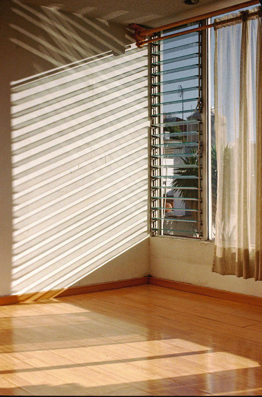 Sunlight streams through a window with blinds, casting shadows on the wall and wooden floor.