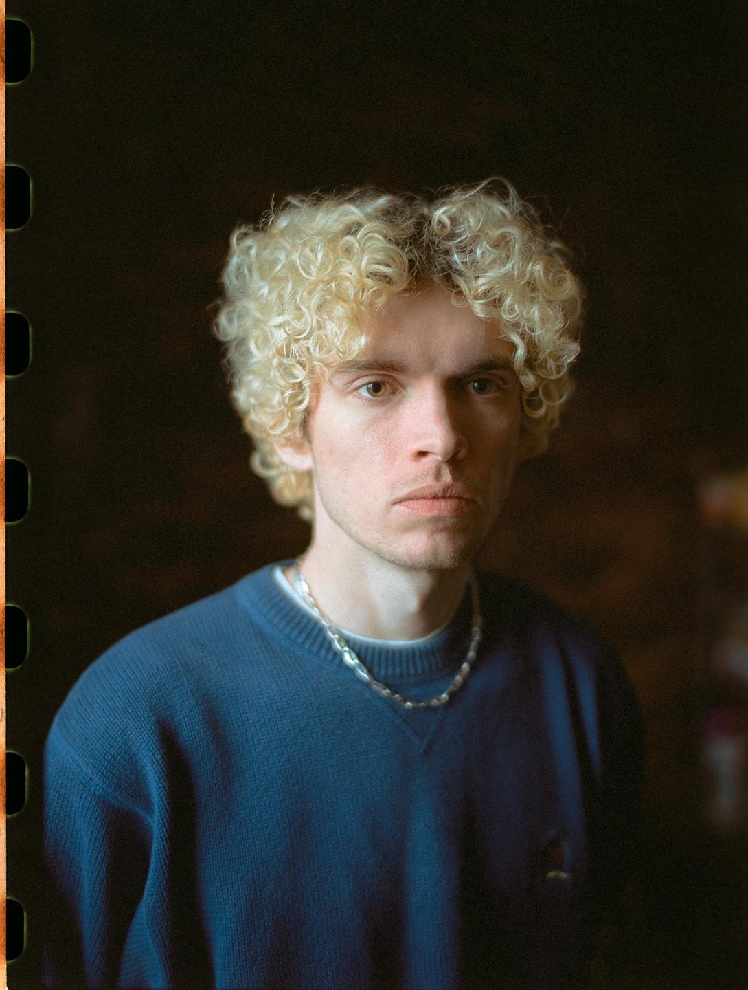 A man with curly blond hair and a blue sweater looks directly at the viewer.