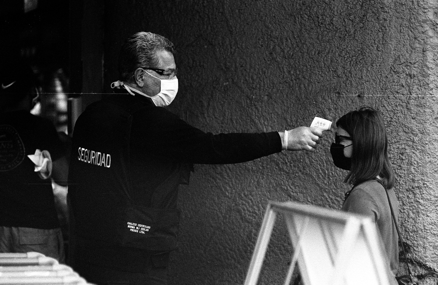 A security guard wearing a face mask and gloves points a thermometer at a woman's forehead.