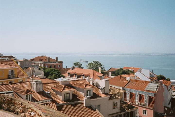 A cargo ship sails on the water behind a city with many buildings with red tile roofs.
