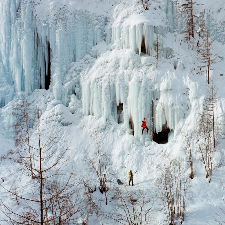 A person in red is ice climbing on a large frozen waterfall while another person stands below in a snowy landscape with bare trees.