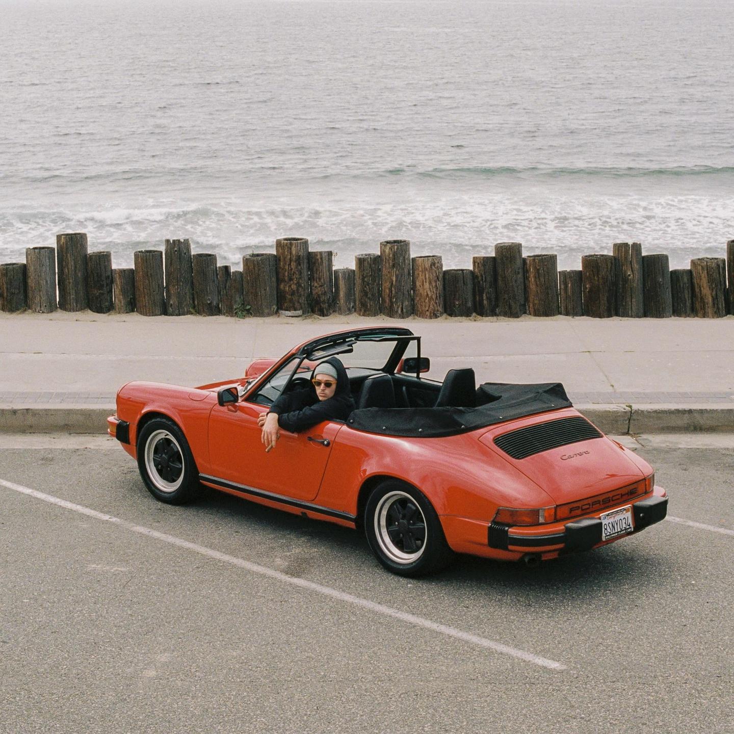 A man in a red convertible car is parked next to a beach with wooden posts.