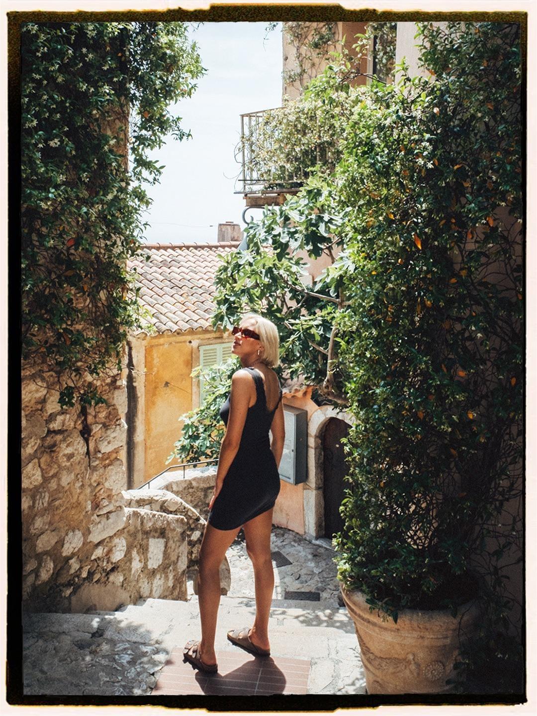 A woman in a black dress stands on stone steps surrounded by lush green foliage and old buildings.