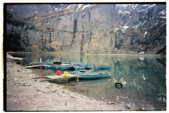 Blue rowboats are docked on a calm lake with a rocky shore and snow-capped mountains in the background.