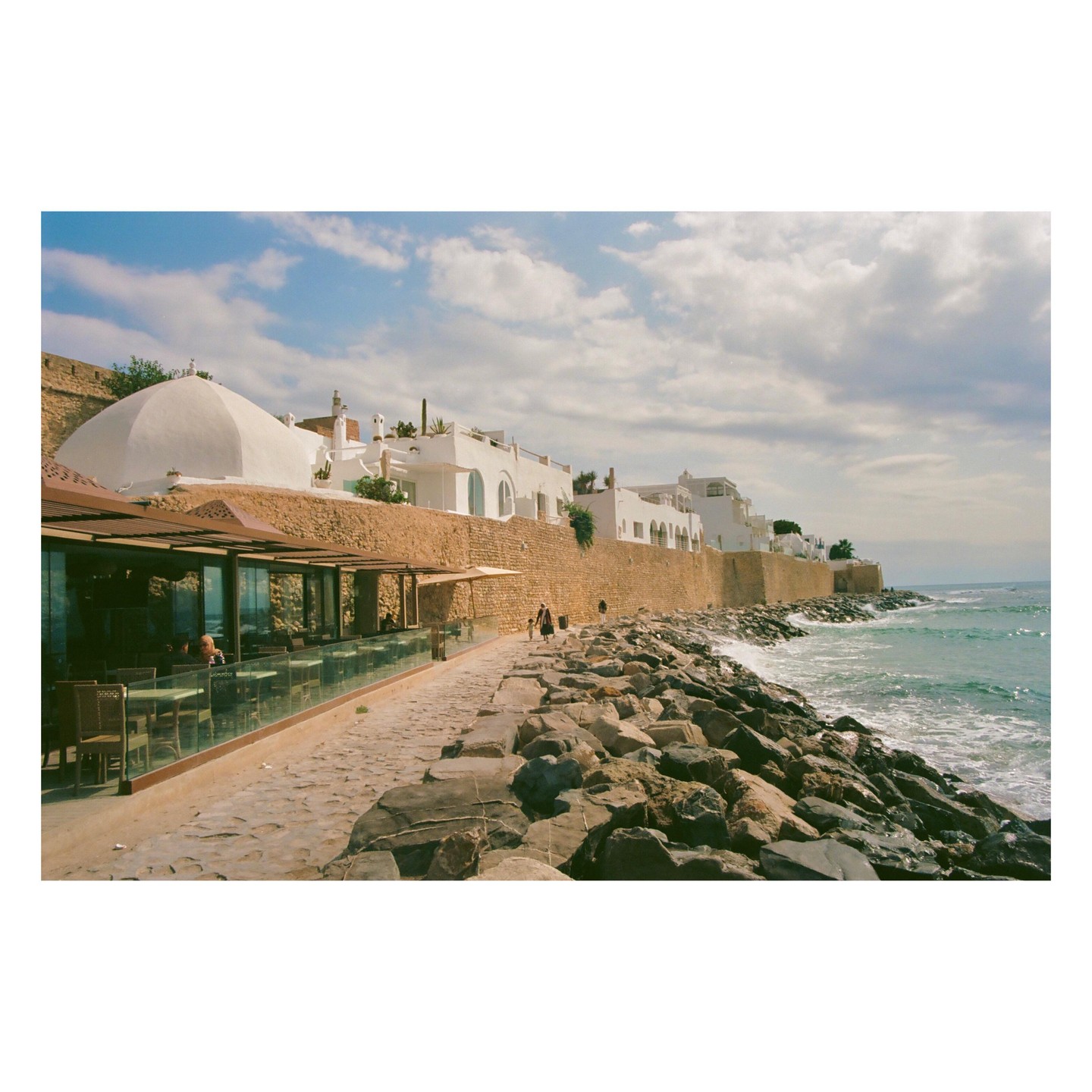 A stone path runs along a rocky coastline with white buildings and a restaurant.