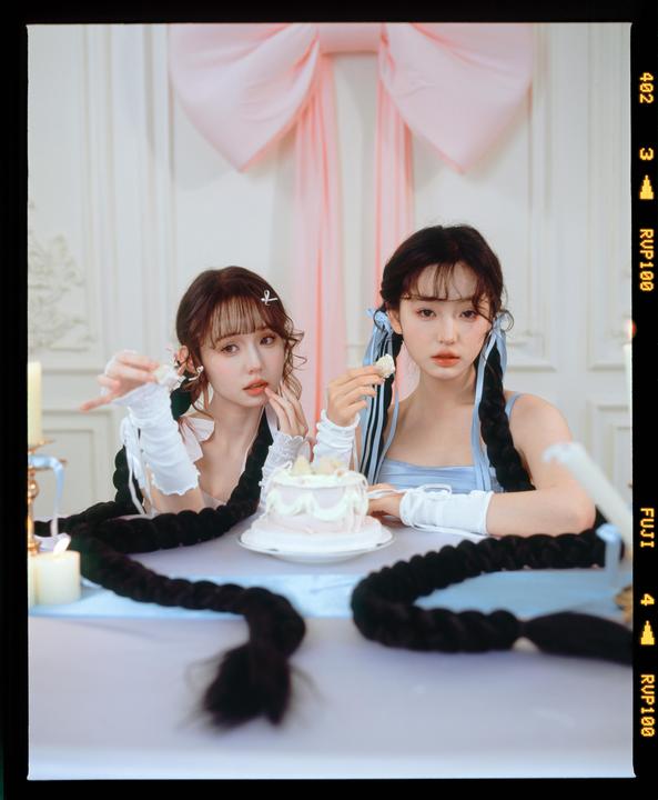 Two women with long braided hair are seated at a table with a small cake and candles, with a large pink bow in the background.