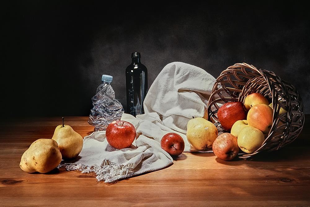 A still life arrangement features a wicker basket spilling apples and pears onto a wooden surface, alongside a crumpled plastic bottle, a glass bottle, and a white cloth.